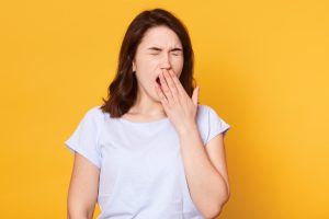 Beautiful woman yawns isolated over yellow background, wears casual white t shirt, has wavy hair, keeps hand near mouth. Free space for advertisment or promotional text. People and lifestyle concept.