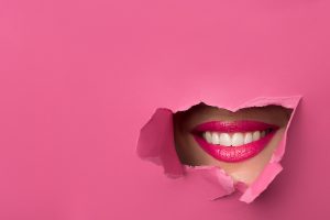 close-up of female lips with pink lipstick and a broad smile that peeps through the pink torn paper