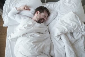 Man in white pajamas has a terrible nightmare, he grinds teeth and prepared a fist for protection, lying in his bed. Concept of insomnia and mental health. Top view.