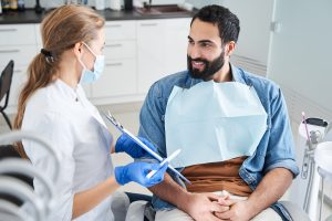 Young female dentist showing the paper to a satisfied smiling bearded man client of the dental office. People talking and looking at each other. People, medicine, stomatology and health care concept