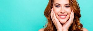 Advertising concept. Portrait of young woman with big toothy smile, modern curly ginger hairdo, enjoying her perfect skin after cream, lotion, peeling isolated on vivid turquoise background