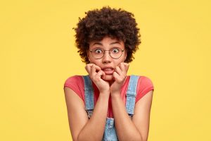 Headshot of frustrated mixed race young woman has crisp hair, bites finger nails with nervous expression, stares with eyes full of fear, wears casual clothes, stands against yellow studio wall