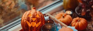 Halloween cozy mood composition on the windowsill. Lighting jack-o-lantern, decorative pumpkins, cones, candles on wooden tray and straw napkin, warm plaid. Hygge halloween home decor. Selective focus.