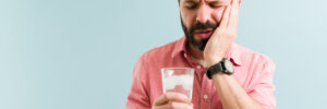 Stressed young man drinking cold water with ice and suffering with tooth sensitivity. Latin man rubbing the side of his face
