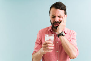 Stressed young man drinking cold water with ice and suffering with tooth sensitivity. Latin man rubbing the side of his face