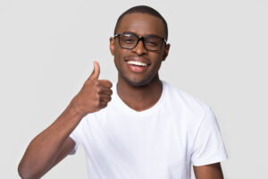 African American man wearing glasses with healthy white smile showing thumbs up, young satisfied client, customer recommend service or product, looking at camera, isolated on studio background