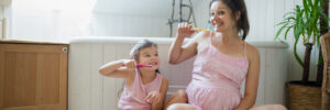Portrait of happy pregnant woman with small daughter indoors in bathroom at home, brushing teeth.