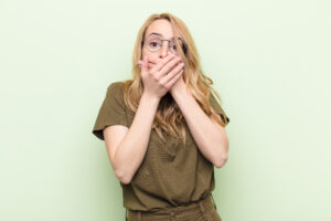 young pretty blonde woman covering mouth with hands with a shocked, surprised expression, keeping a secret or saying oops against flat color wall