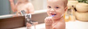 a little boyl with short hair sitting at home in the bathroom on the sink and brushing his teeth with a large blue toothbrush.