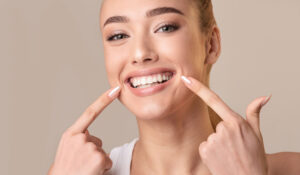 Perfect Smile. Young Woman With White Teeth Touching Mouth Smiling To Camera Posing On Beige Background. Studio Shot