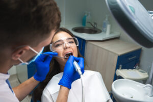 dentist in mask filling the patient's root canal while she is lying on dental chair wearing safety glasses under the medical lamp in clinic.