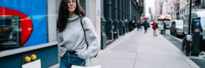 Cheerful relaxed female in sweater and jeans walking on sidewalk with shopping bags and looking dreamily away in New York