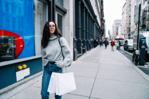 Cheerful relaxed female in sweater and jeans walking on sidewalk with shopping bags and looking dreamily away in New York