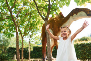 Cheerful little girl having fun in park with dinosaur replicas outdoors.