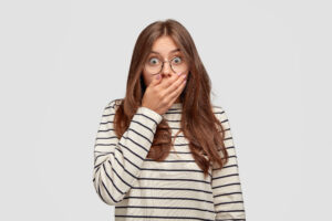 Horizontal shot of stressful stratled young woman gasps from shock and fear, keeps hand on mouth, stares with bugged eyes, wears casual oversized striped sweater, isolated over white background