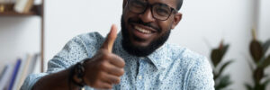 Head shot positive confident black businessman sitting at office desk smiling looking at camera showing hand gesture thumbs up sign symbol of good result, recommendation and success in work or study