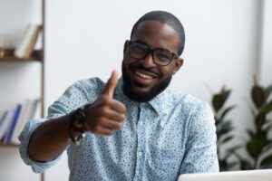 Head shot positive confident black businessman sitting at office desk smiling looking at camera showing hand gesture thumbs up sign symbol of good result, recommendation and success in work or study