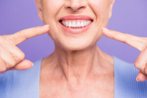 Concept of having strong healthy straight white perfect teeth at old age. Cropped portrait of beaming smile female pensioner pointing on her teeth, isolated over violet background
